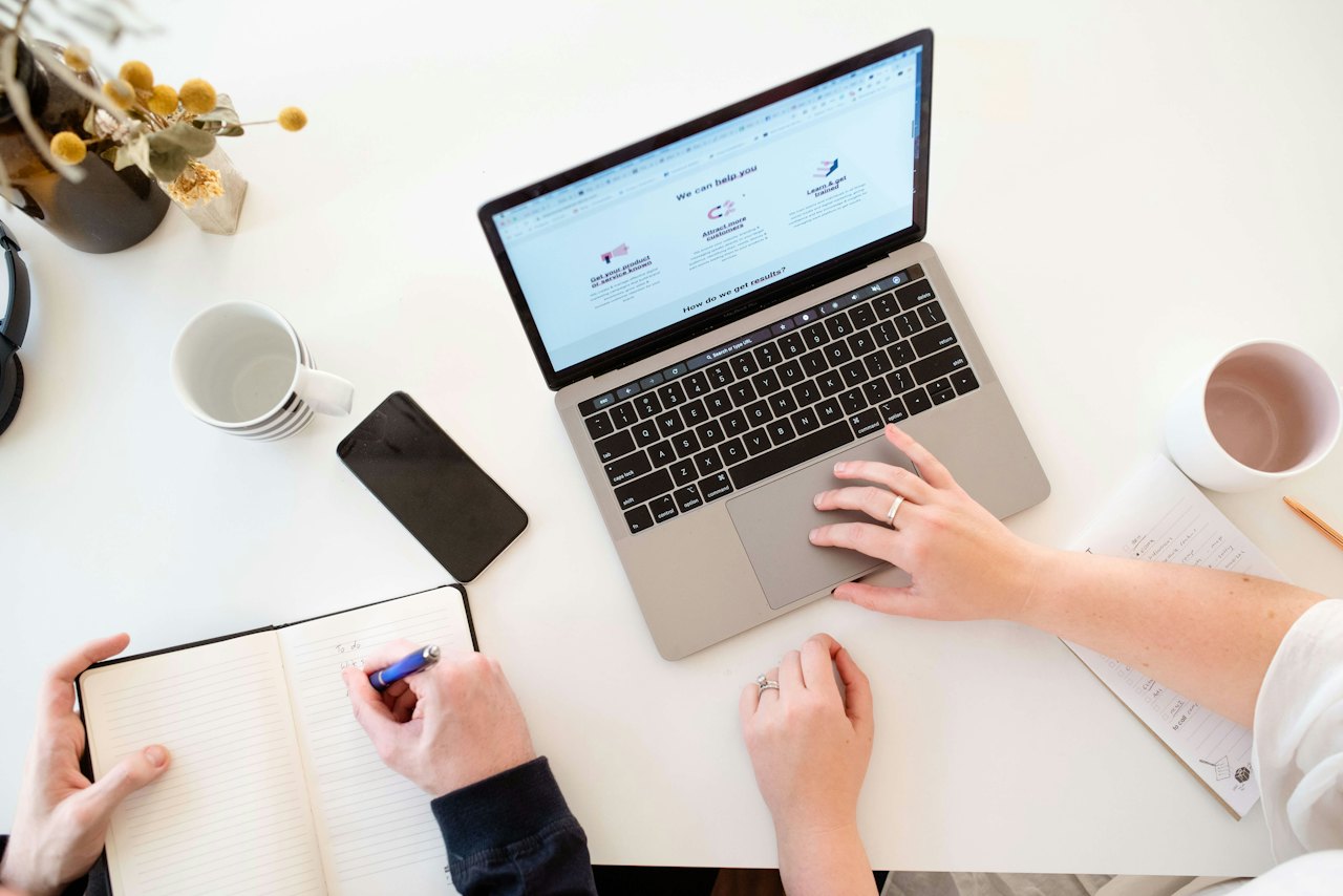 Vista cenital de una mesa de trabajo: dos personas con un portátil, una libreta y café.